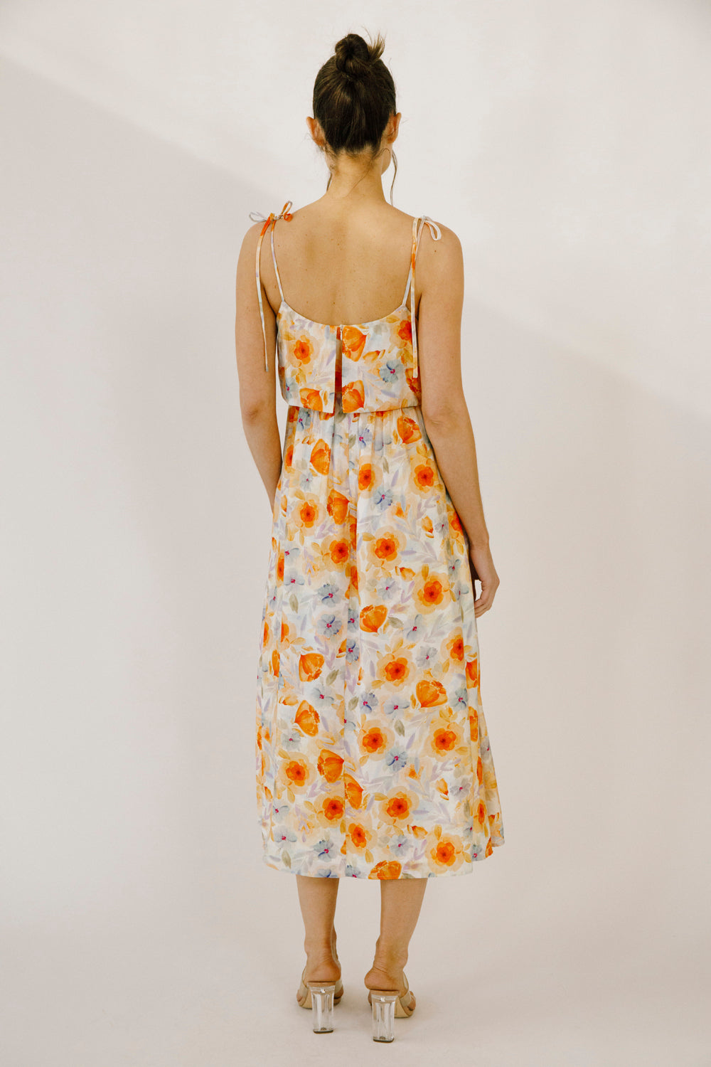 A woman viewed from behind wearing a colorful floral print midi dress with a soft watercolor effect, self-tie straps and an invisible zipper against a neutral wall.