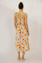 A woman viewed from behind wearing a colorful floral print midi dress with a soft watercolor effect, self-tie straps and an invisible zipper against a neutral wall.