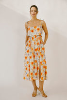 Model wearing a colorful floral print midi dress with a soft watercolor effect, self-tie straps, side pockets and a leg slit against a neutral wall.