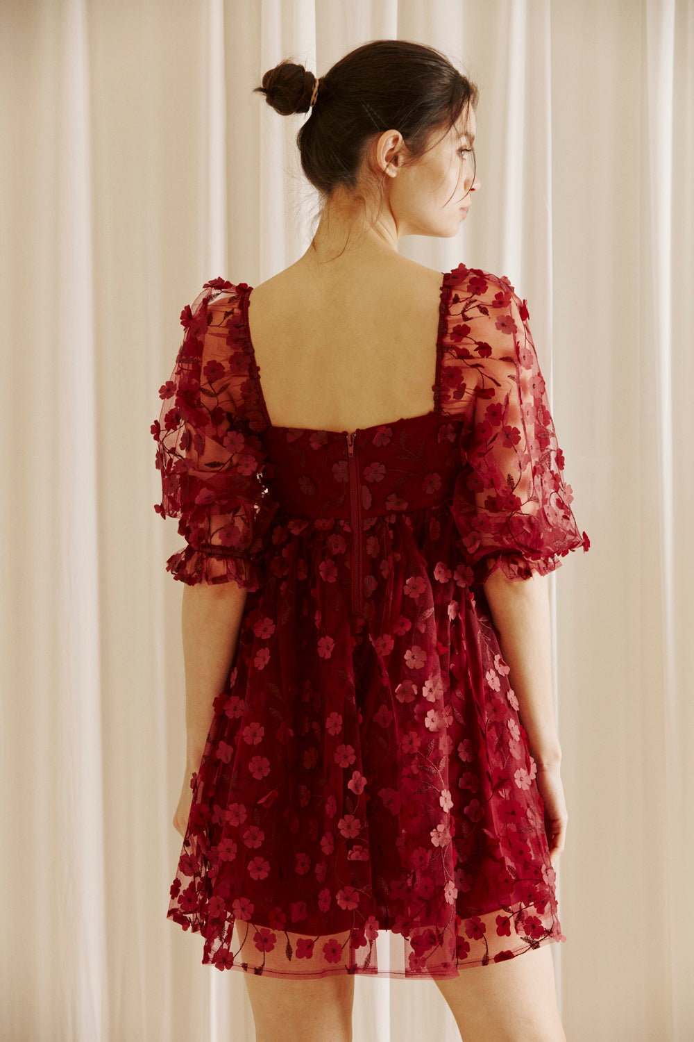 Back view of a woman wearing a burgundy dress with 3D floral detail throughout against a white curtain background