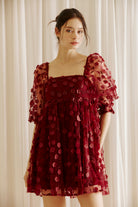 Woman wearing a burgundy dress with 3D floral detail throughout against a white curtain background