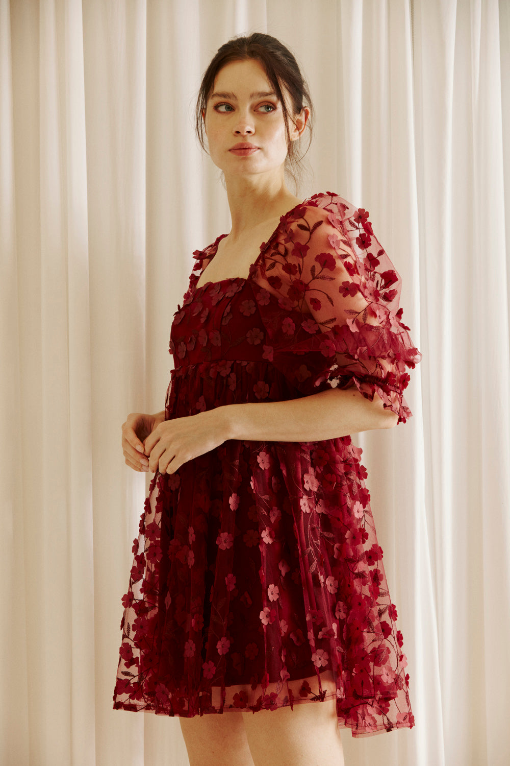 Side view of a woman wearing a burgundy dress with 3D floral detail throughout against a white curtain background