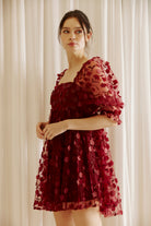 Side view of a woman wearing a burgundy dress with 3D floral detail throughout against a white curtain background