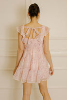 Back view of a woman wearing a pink dress with a cherry blossom print, self-tie bow and smocked upper back against a neutral background
