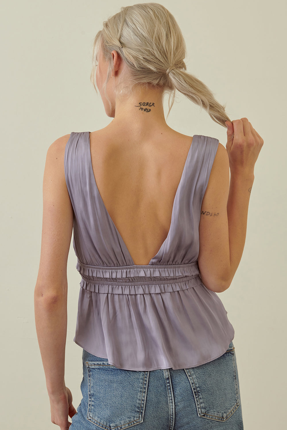 A woman shown from the back wearing a sleeveless lavender top with an open back and gathered waist, paired with blue jeans against a plain light beige background.