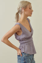 A woman wearing a lavender sleeveless peplum top with a gathered waist, paired with blue jeans, shown from a side profile against a plain beige background.