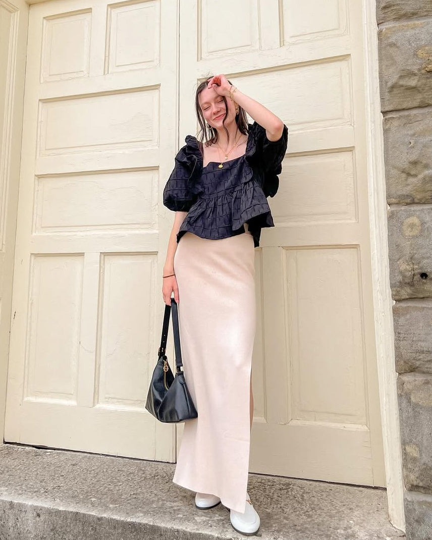 Woman wearing a black blouse and cream skirt posing in front of a large door with stone walls.