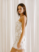 Side view of a woman wearing a white 3D floral embroidery mini dress against a white curtain background