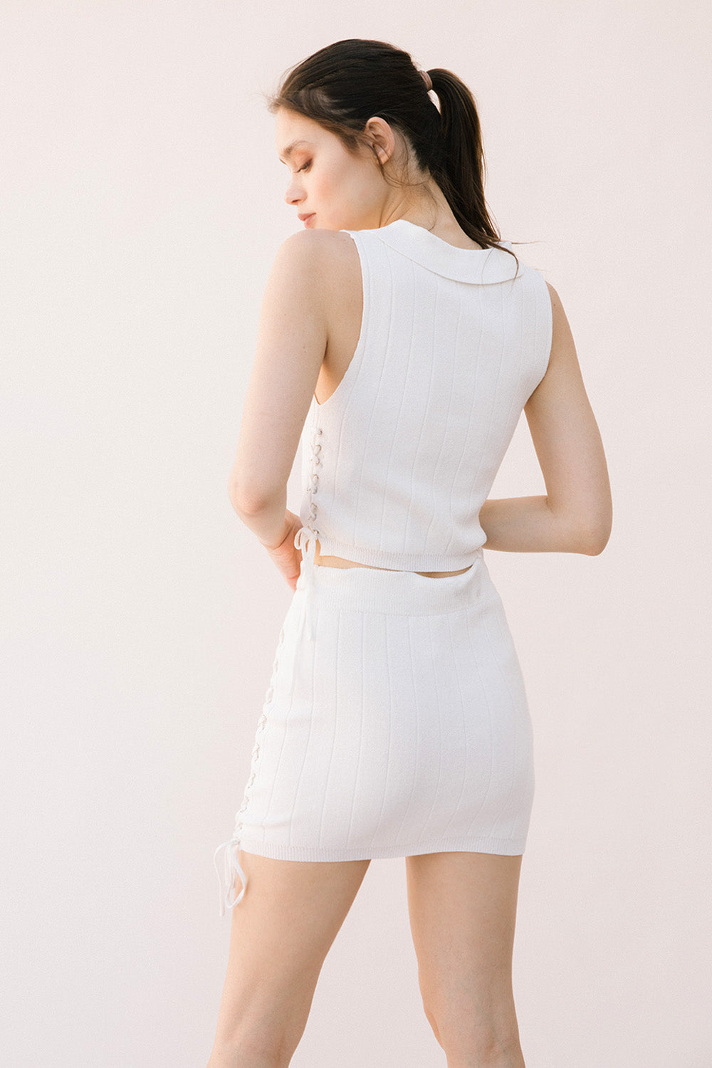 Back view of a woman wearing a white sleeveless top and skirt set on a light background