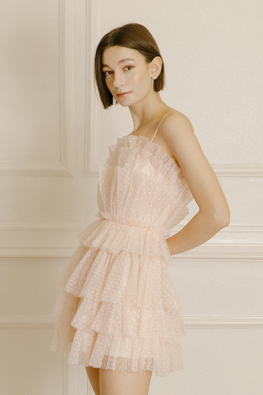 Side of a woman wearing a soft pink mini dress with white dots and tiered tulle on a beige background