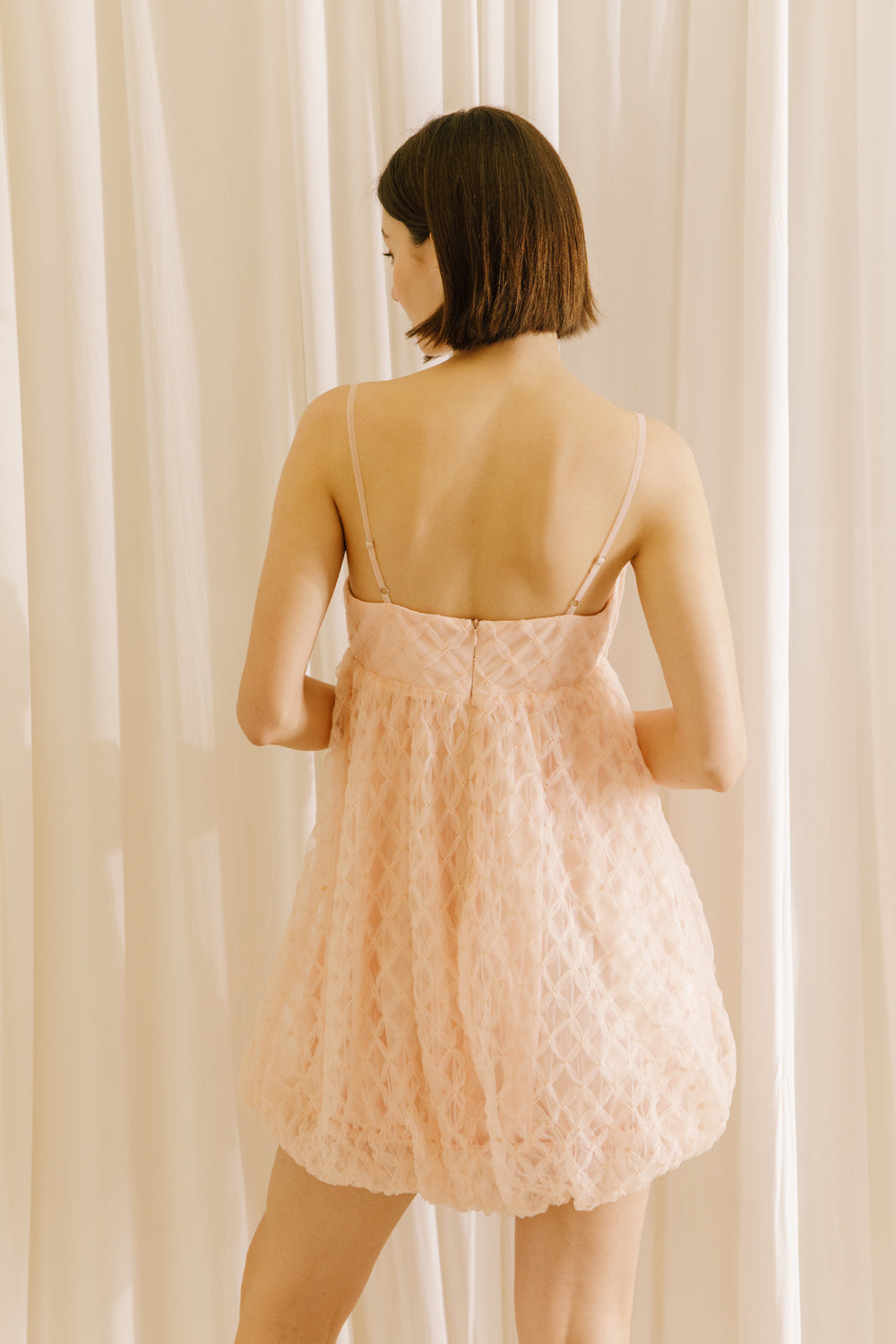 Back view of a woman wearing a light pink dress with a voluminous bubble-hem against a beige curtain background