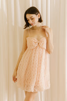 Front view of a woman wearing a light pink dress with a voluminous bubble-hem against a beige curtain background