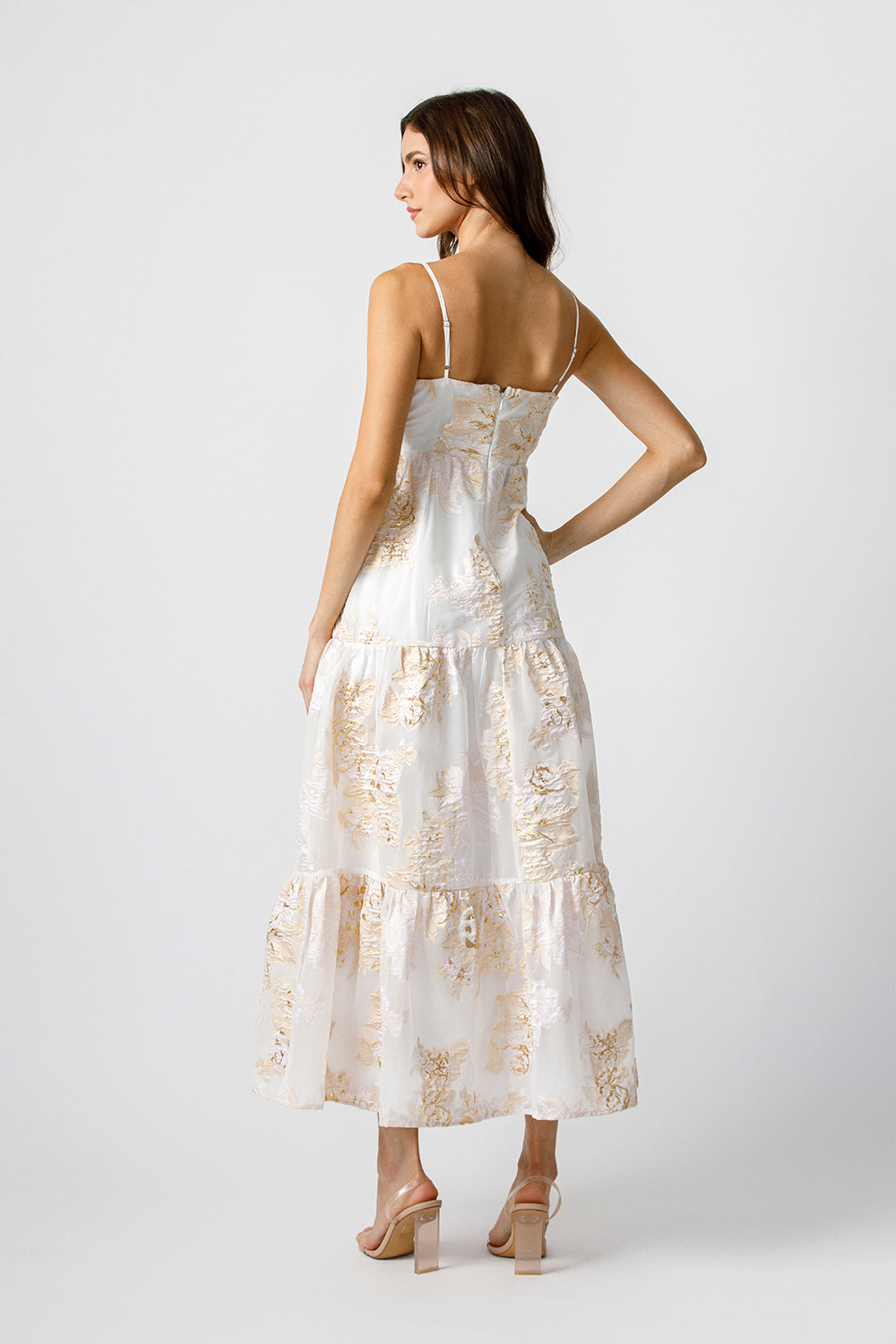 Back view of a woman wearing a white dress with gold floral pattern on a white background