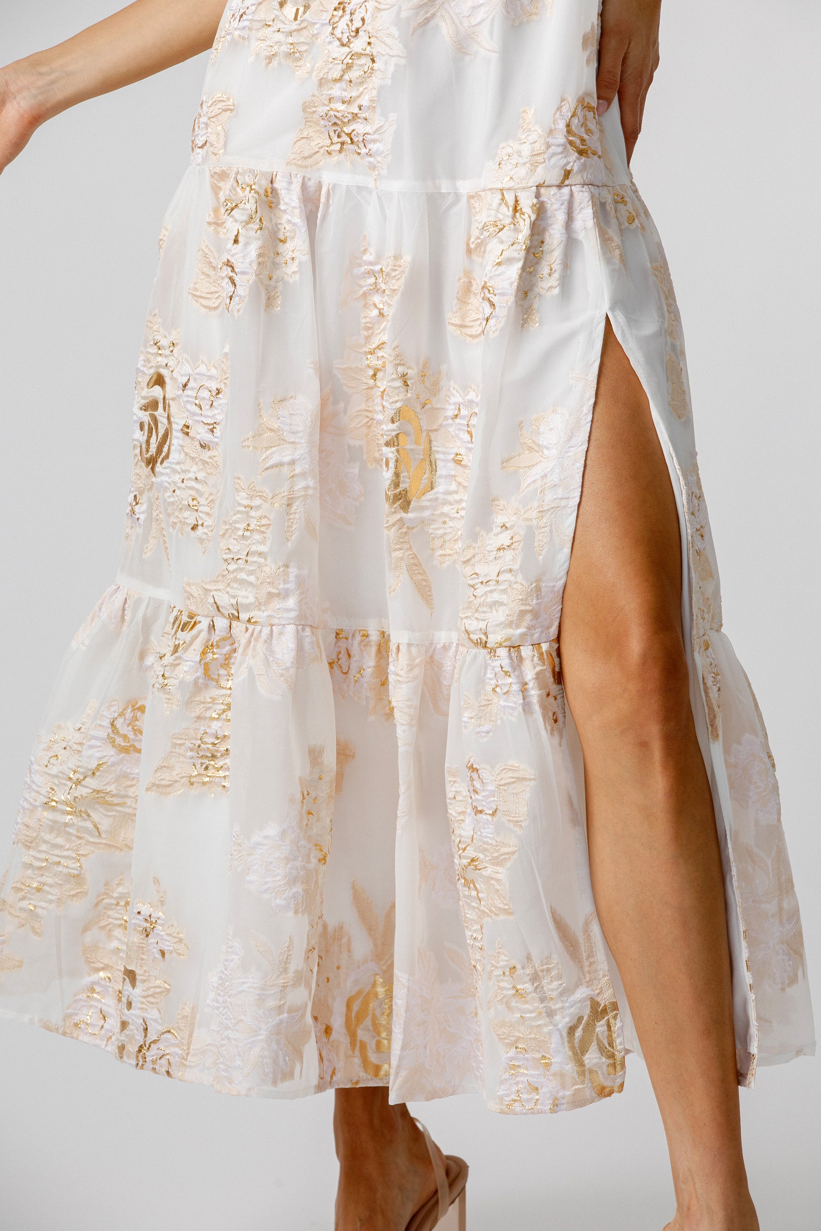 Details of a white dress with gold floral pattern and a slit on a plain background