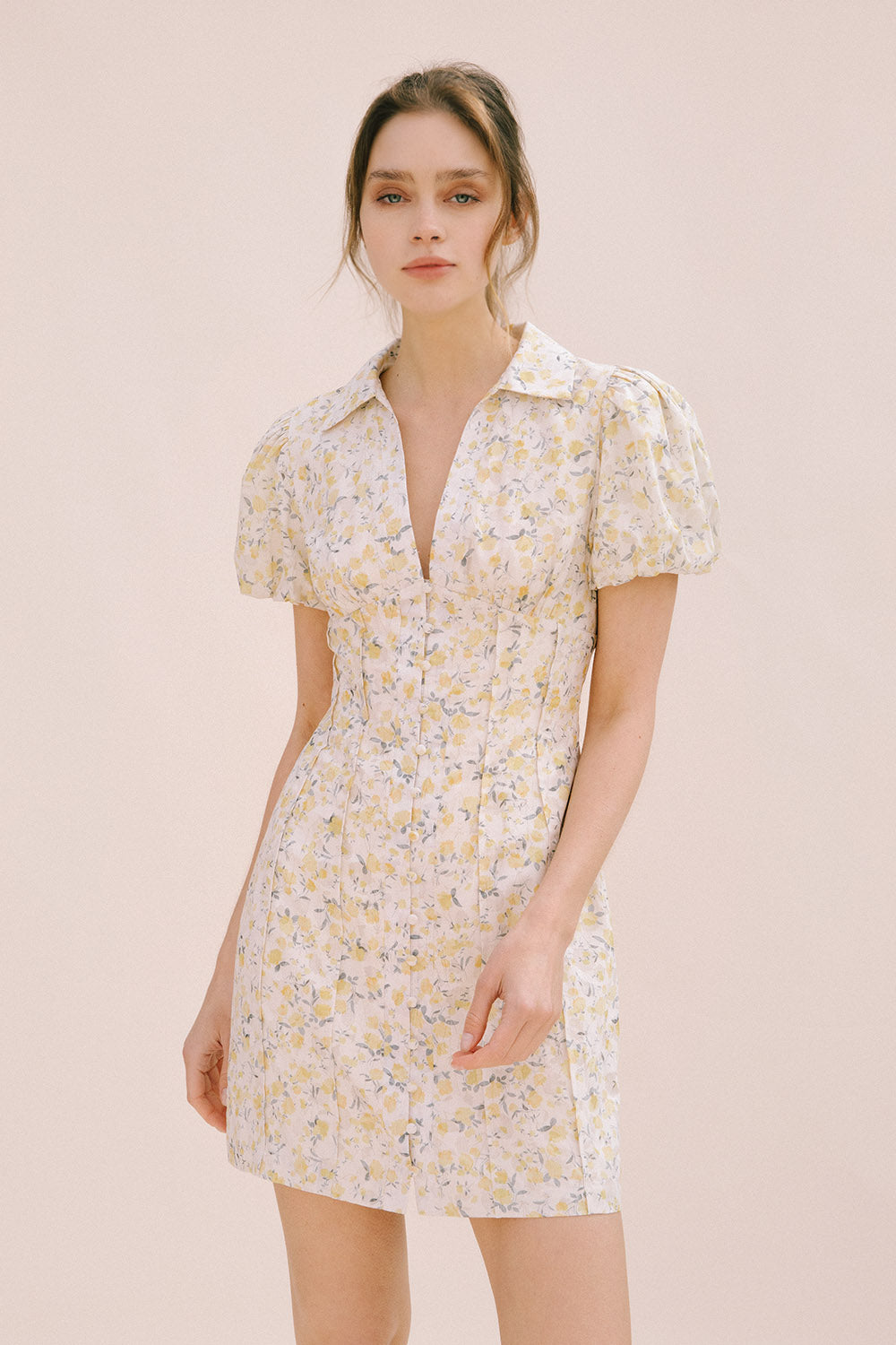 Model wearing a cream dress with a floral pattern, center buttons, a bodycon fitted body and vertical darts from the empire waist down the dress (creating a corset-like detail).