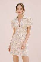 Model wearing a cream dress with a floral pattern, center buttons, a bodycon fitted body and vertical darts from the empire waist down the dress (creating a corset-like detail).