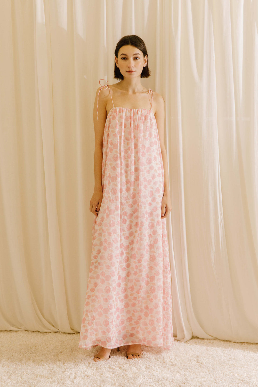 Woman wearing a pink tulip floral dress standing against a beige curtain.