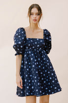 Woman wearing a navy blue dress with puff sleeves and floral pattern on a beige background