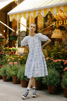 Woman in a blue checkered dress standing in front of a store with flowers and plants.