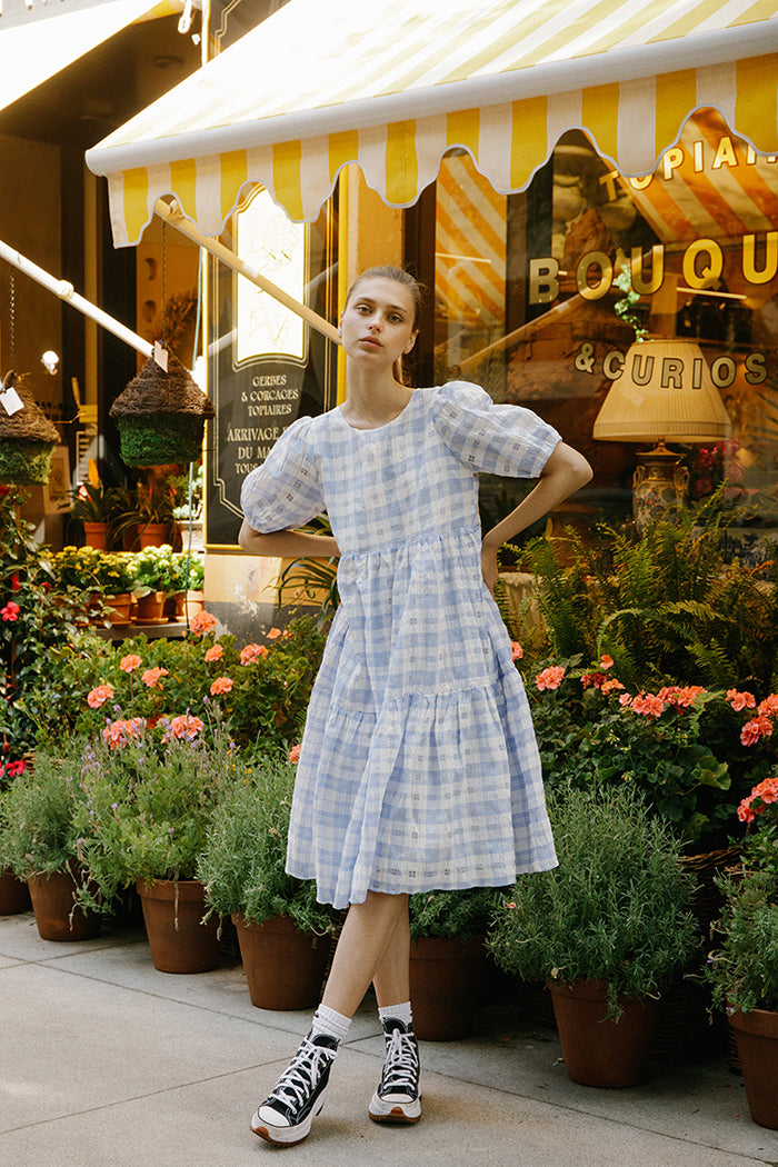 Woman in a blue checkered dress standing in front of a store with flowers and plants.