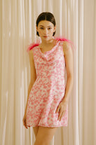 Woman wearing a white dress with a fun pink pattern, cowl neckline and straps adorned with pink feathers against a beige curtain background