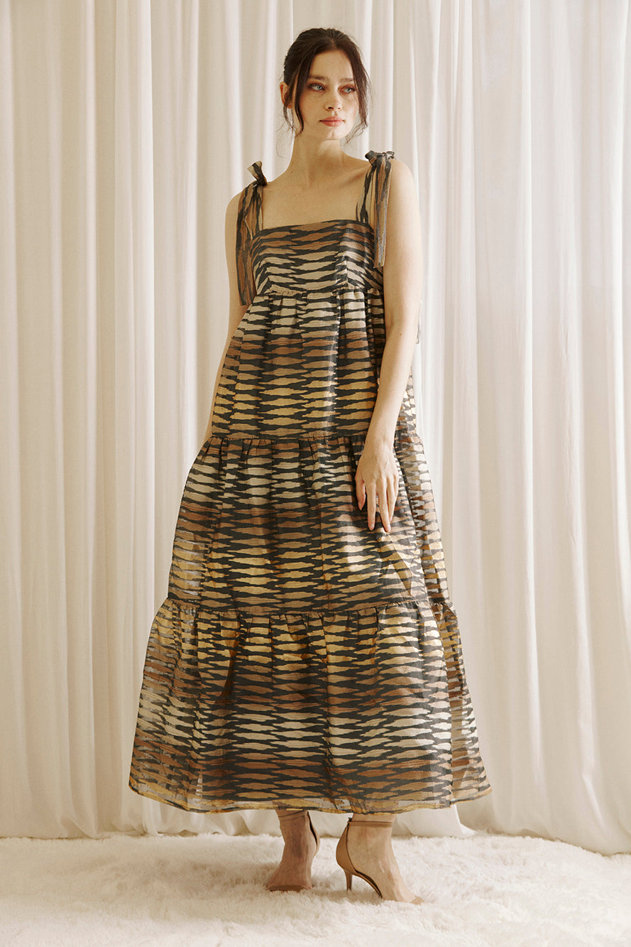 Woman wearing a brown and gold patterned dress standing against a white curtain.