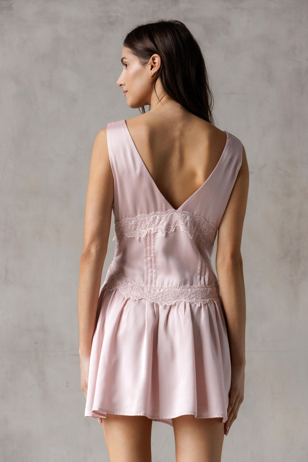 Back of a woman wearing a pink dress  with lace trim detailing against a plain 