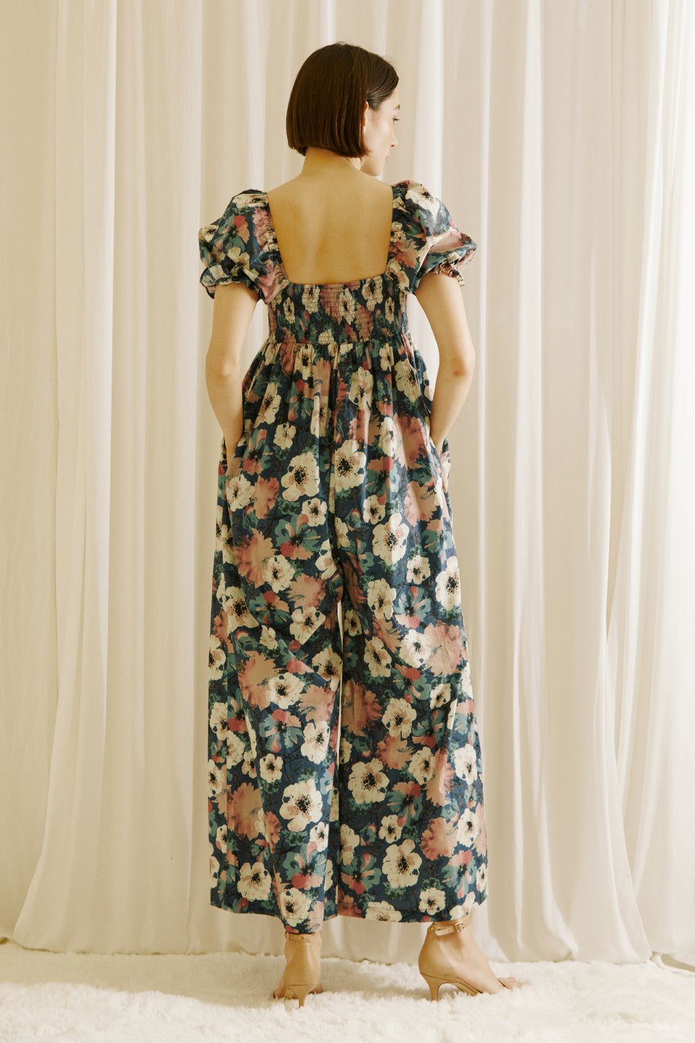 A woman viewed from behind wearing a flowy jumpsuit standing in front of white curtains