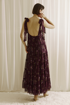 A woman viewed from behind wearing a plum base maxi dress with an allover pink floral print and a low scoop open back on a white background
