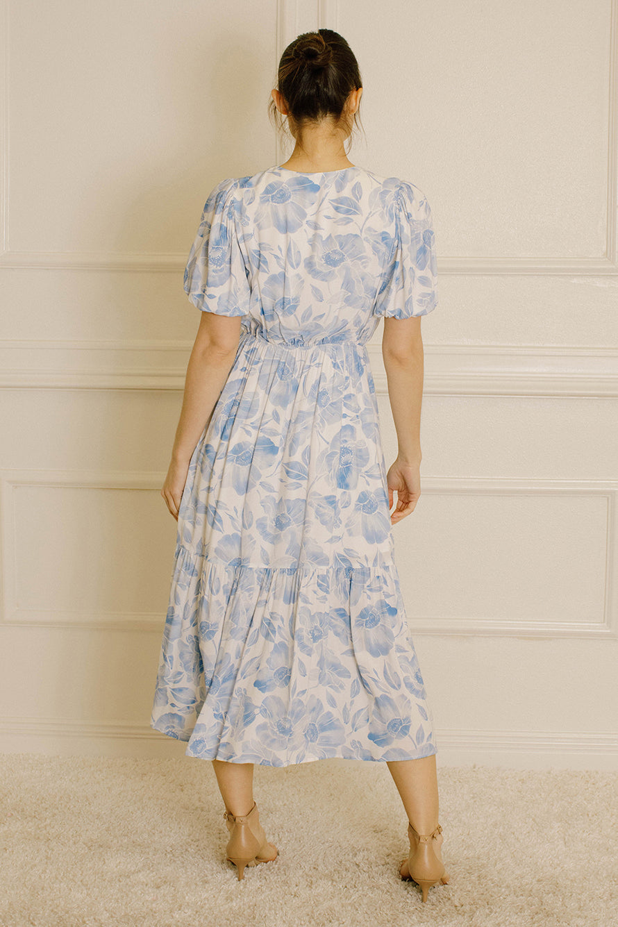 A woman viewed from behind wearing a light blue and white floral print midi dress with puff sleeves and a tiered skirt, standing against a cream paneled wall.