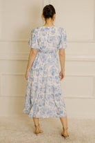 A woman viewed from behind wearing a light blue and white floral print midi dress with puff sleeves and a tiered skirt, standing against a cream paneled wall.