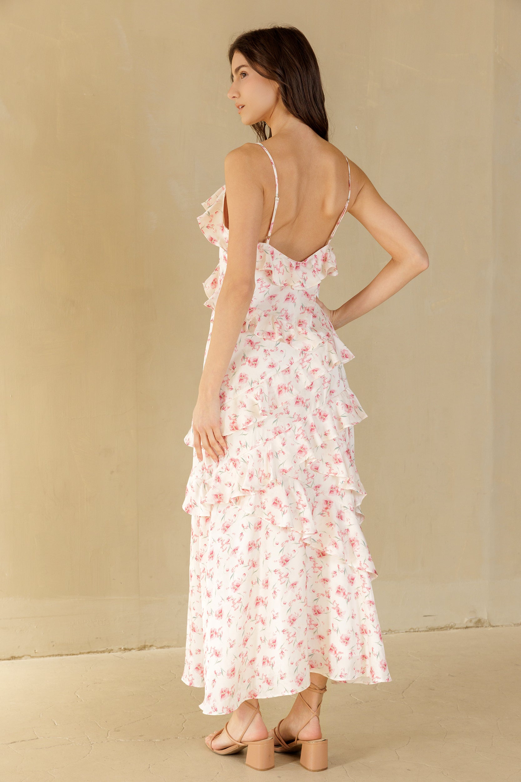 A woman modeling a white tiered maxi dress with a blush floral print, shown from the back against a neutral beige background.