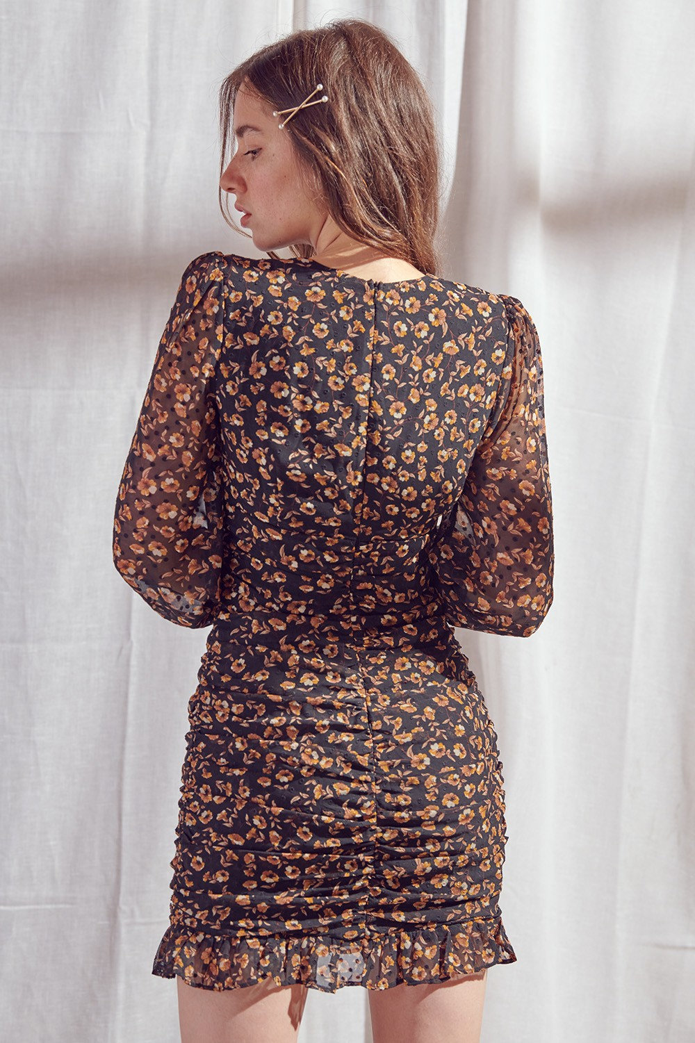 A woman wearing a ruched black mini dress with a orange and yellow floral print, photographed from the back against a white curtain backdrop.