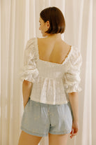 A woman shown from the back wearing a white puff-sleeve lace top with a square neckline and ruffled trim, paired with light blue denim shorts, standing in front of cream-colored curtains.