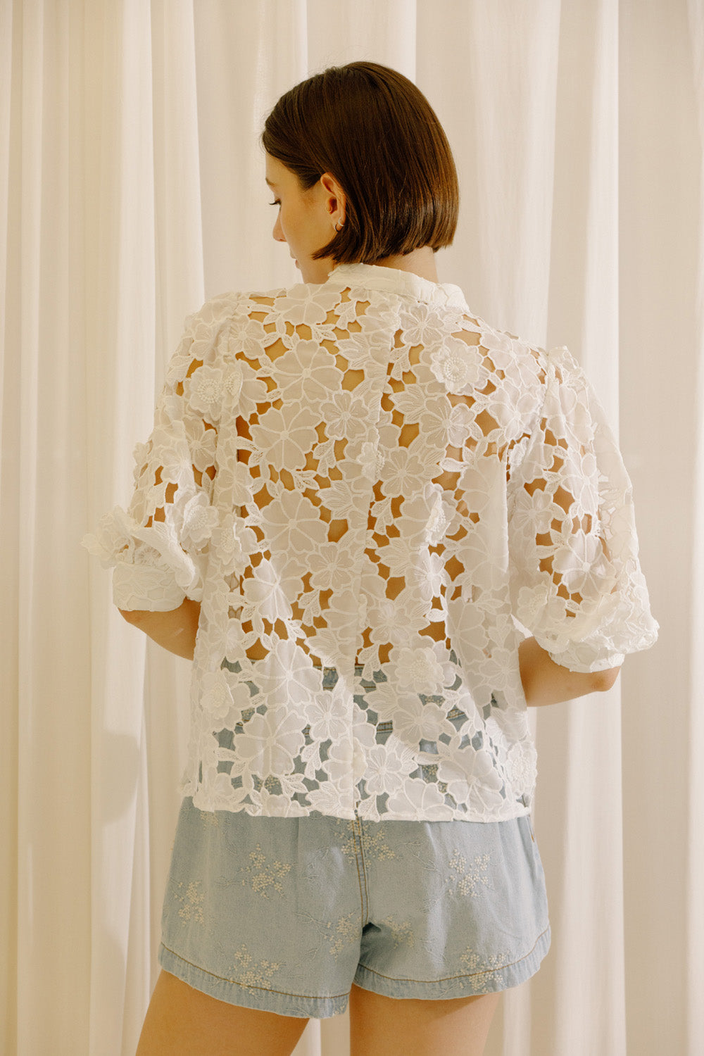 A woman viewed from behind wearing a white eyelet short-sleeve blouse and light blue denim shorts, standing in front of white curtains.