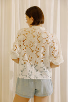 A woman viewed from behind wearing a white eyelet short-sleeve blouse and light blue denim shorts, standing in front of white curtains.