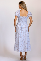 Back view of a woman wearing a blue base midi dress with pink florals, a fully smocked bodice, puff sleeves, and a flowy midi skirt.