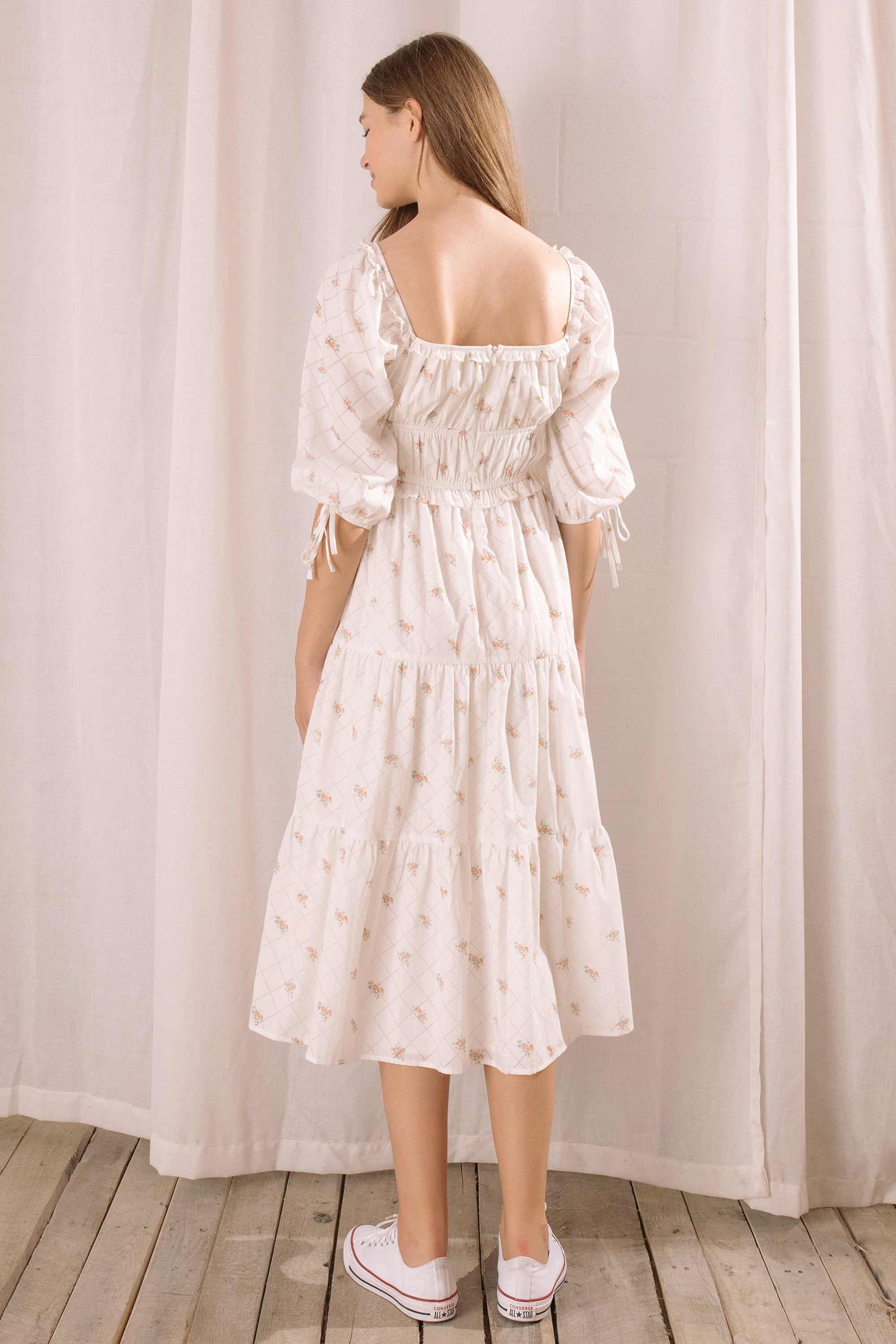 A woman viewed from behind wearing a white dress with puff sleeves against a neutral curtain background