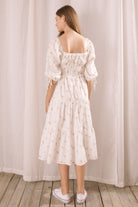 A woman viewed from behind wearing a white dress with puff sleeves against a neutral curtain background
