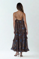 Back view of a model wearing a flowy brown maxi dress with blue floral print and cascading ruffle tiers for an effortless flowy look.