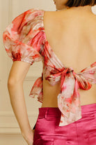 A woman shown from the back wearing a sheer puff-sleeve top with a pink, red, and white watercolor floral print and a large tie-back bow. She is also wearing bright pink satin pants.