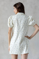 A woman shown from the back wearing a short puff-sleeve collared dress with a small blue floral print on a warm off-white background, set against a textured gray wall.