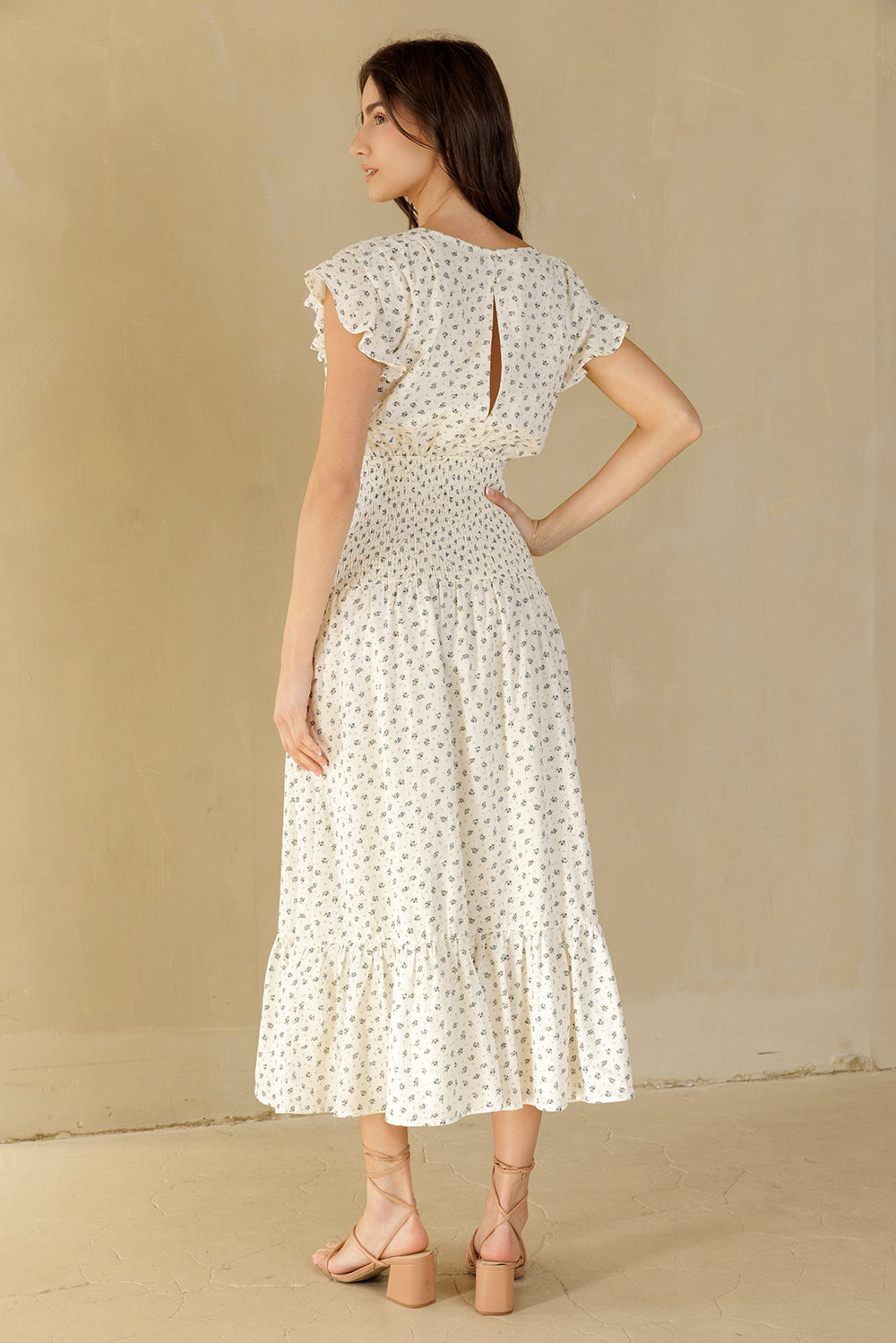 A woman wearing a cream midi dress with a small blue and green floral pattern, shown from the back. The dress features ruffled cap sleeves, a keyhole back with a decorative tie, and a tiered skirt.