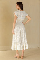 A woman wearing a cream midi dress with a small blue and green floral pattern, shown from the back. The dress features ruffled cap sleeves, a keyhole back with a decorative tie, and a tiered skirt.