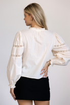 A woman viewed from behind wearing a cream long sleeve top featuring textured Swiss dot cotton, delicate lace paneling, puff sleeves.