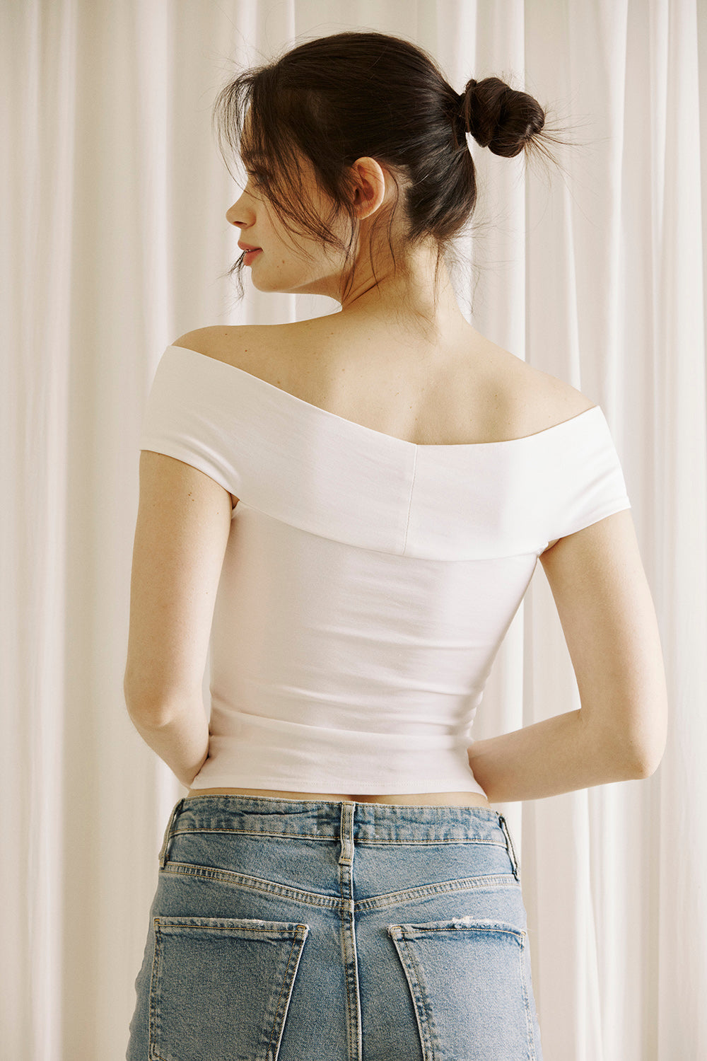 A woman viewed from behind wearing a  white off-shoulder top and blue jeans against a plain background