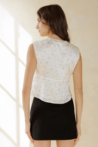A woman shown from the back wearing a sleeveless white top with a very subtle light blue floral pattern and a black mini skirt, set against a warm beige background with soft window light shadows.