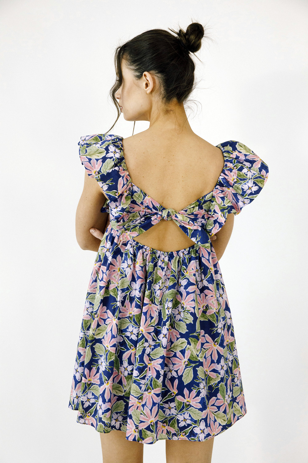 A woman shown from the back wearing a navy blue floral dress with puff sleeves and a large bow detail at the open back, photographed against a plain white studio background.