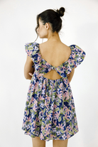 A woman shown from the back wearing a navy blue floral dress with puff sleeves and a large bow detail at the open back, photographed against a plain white studio background.