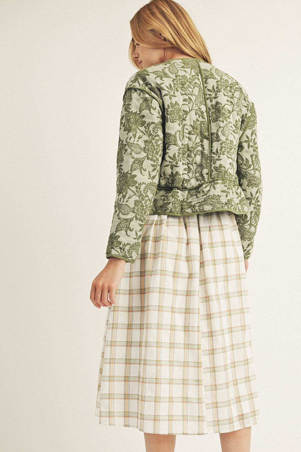 Back of a woman wearing a green floral jacket and plaid skirt on a plain background